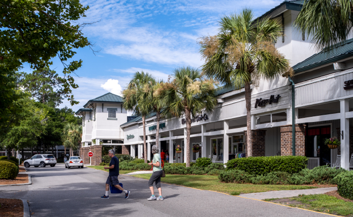 Man and woman exit shops with palm trees and cars in background.