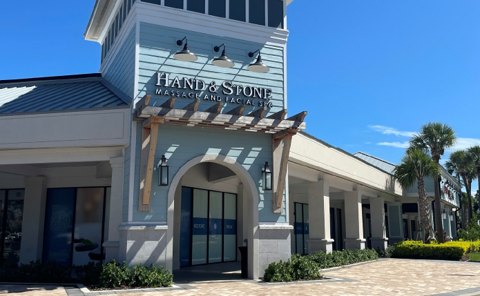 Store front of Hand & Stone with palm trees behind it.