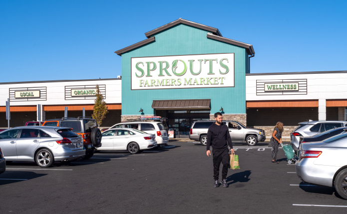 Man with bag in front of Sprouts supermarket in parking lot.