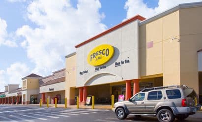 Fresco y Más at Shops at Palm Lakes in Miami, FL, featuring a yellow and cream façade with an arched entryway, bold oval signage, yellow safety bollards, a striped pedestrian crossing, and vehicles entering the front drive aisle beneath bright tropical daylight.