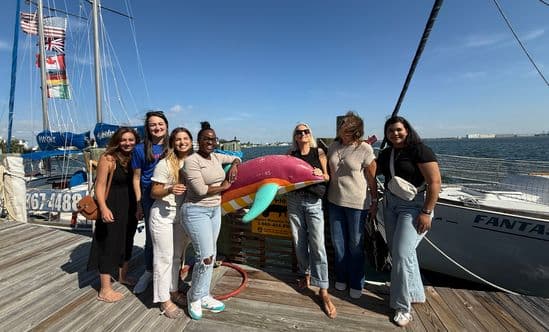 Group of women standing on a waterfront dock beside sailboats, posing with a colorful dolphin sculpture under a clear blue sky, with water and distant shoreline in the background.