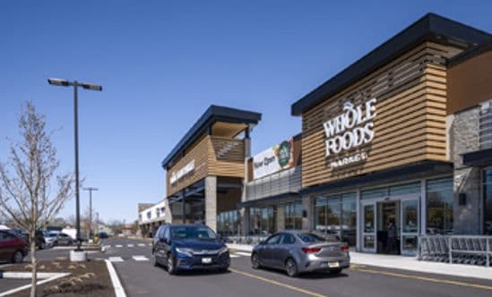 Two cars driving in front of Whole Foods Market storefront at Barn Plaza in Doylestown, PA, featuring modern architectural design, active customer traffic, and a newly paved parking area within the retail center.