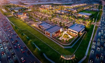 Aerial twilight rendering of Block 59 in Naperville, IL, showcasing the full mixed‑use development with multiple restaurant and retail buildings, illuminated pedestrian areas, landscaped open spaces, extensive parking fields, and adjacent highway traffic flowing along the site’s frontage.