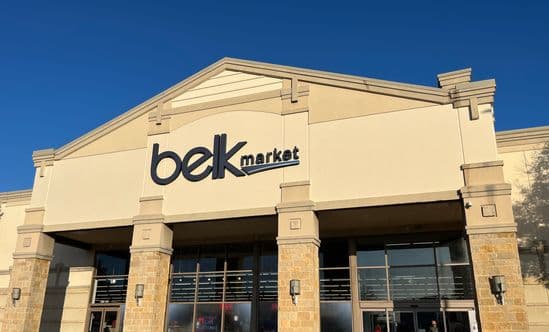 Belk Market storefront featuring a beige and stone façade with large glass windows and entrance doors, illuminated by sunlight under a clear blue sky.