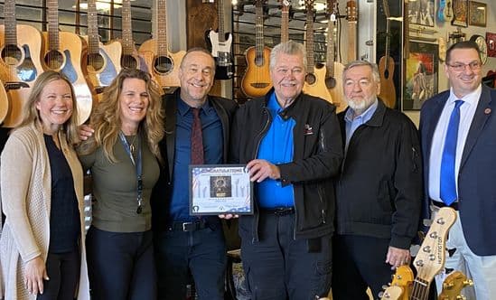 Group of six people posing in a guitar shop, with one holding an “Organization of the Month” certificate in front of a wall of guitars.