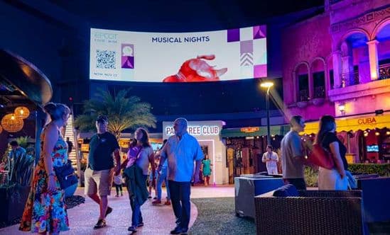 People walking in a shopping center courtyard at night with an oversized digital screen showing behind them