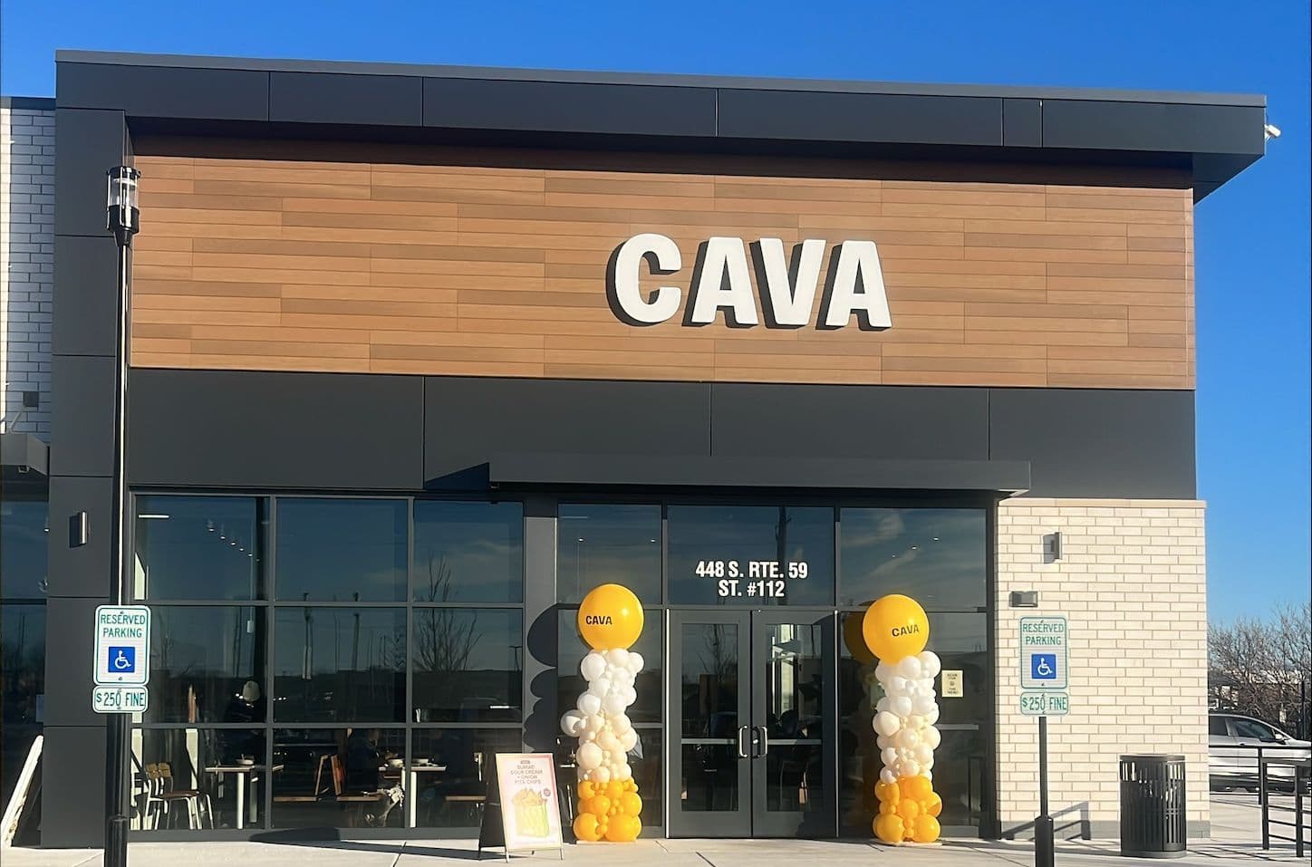 Front exterior of a CAVA restaurant at Block 59 at Westridge Court in Naperville, Illinois, with a one‑story gray and wood façade, large white “CAVA” sign above glass entrance doors, balloon columns on each side of the doorway, and a paved walkway and parking area in front beneath a clear blue sky