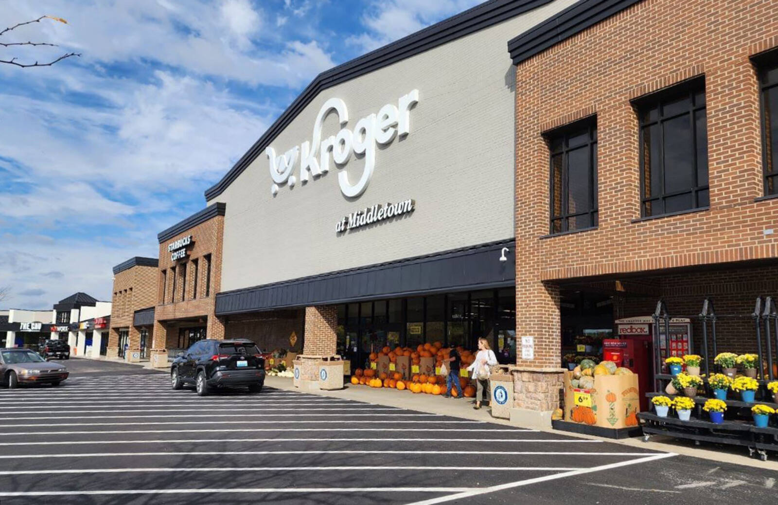 Kroger store entrance with shoppers in entrance and 2 cars on access road in front.