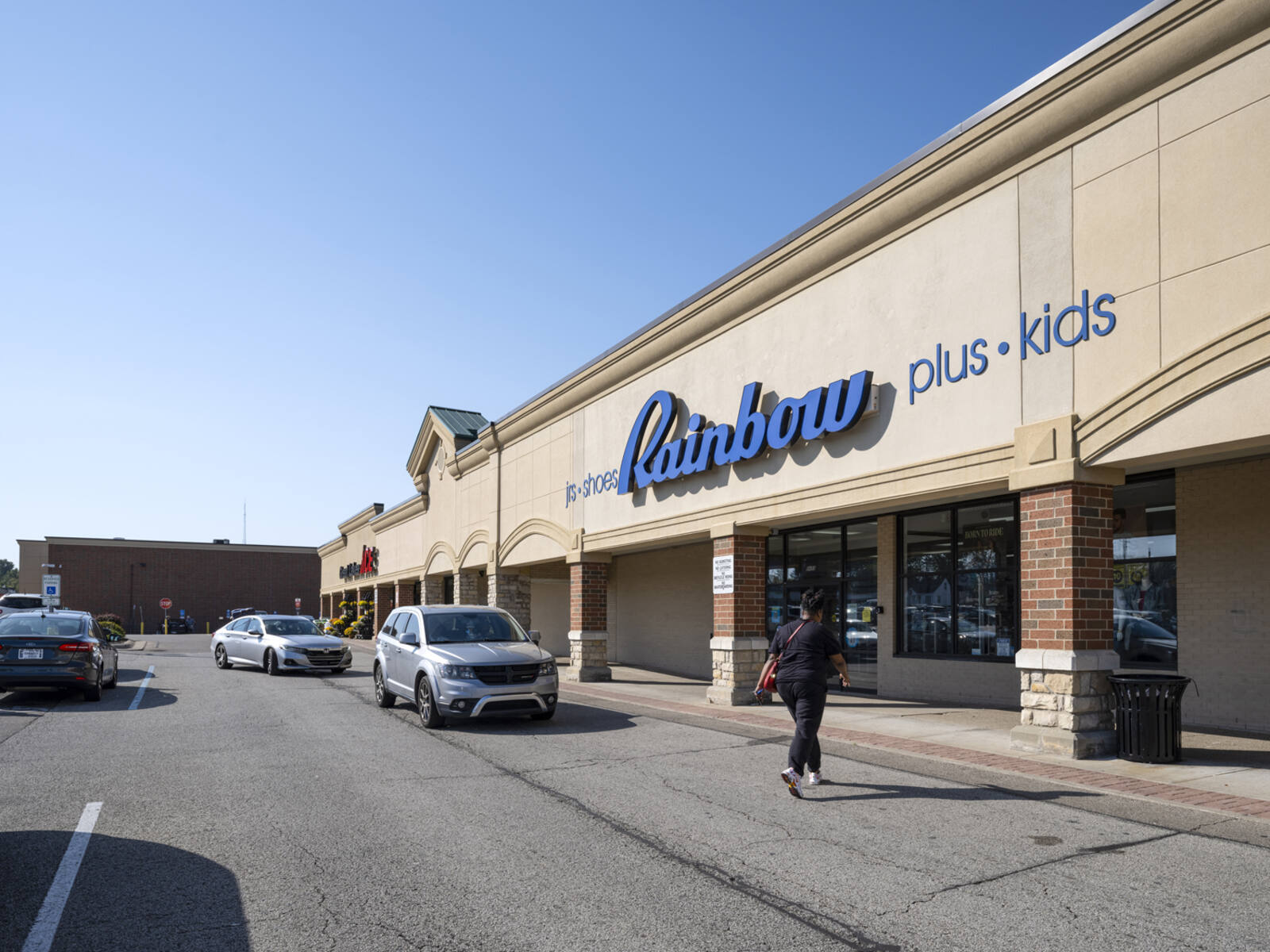 Patron crosses sidewalk at Rainbow store with cars stopped in crosswalk.