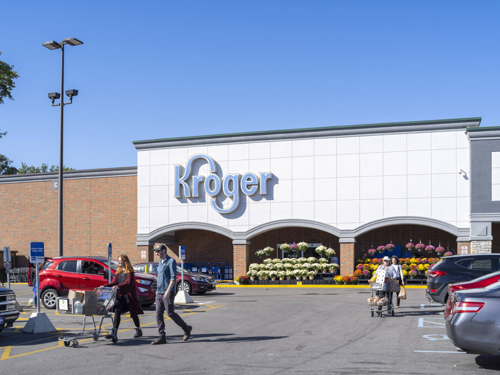 People with shopping carts walking back to their cars in front of Kroger that is lined with potted flowers
