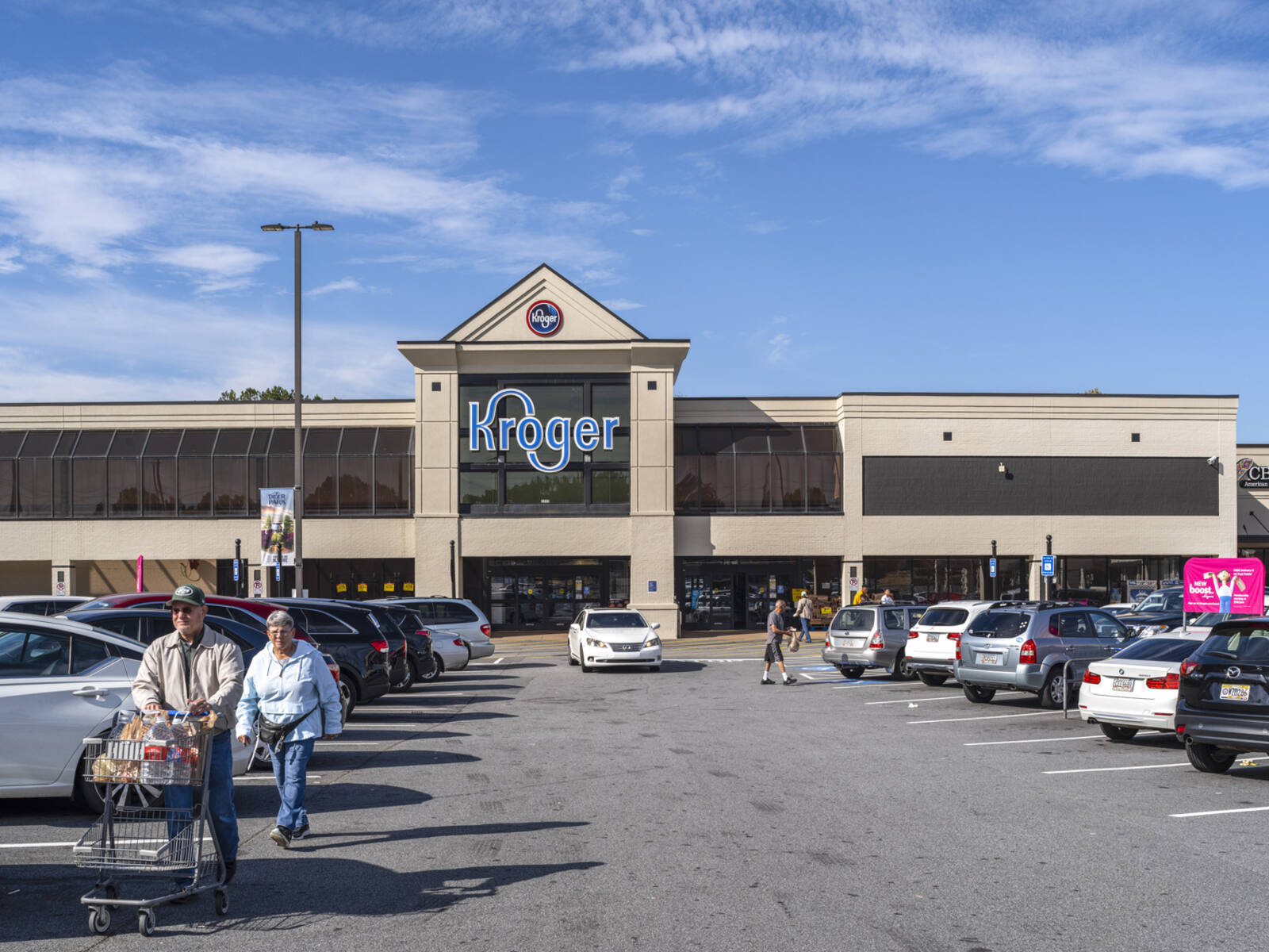 Customers with carts walking up parking aisle in front of Kroger grocery store.