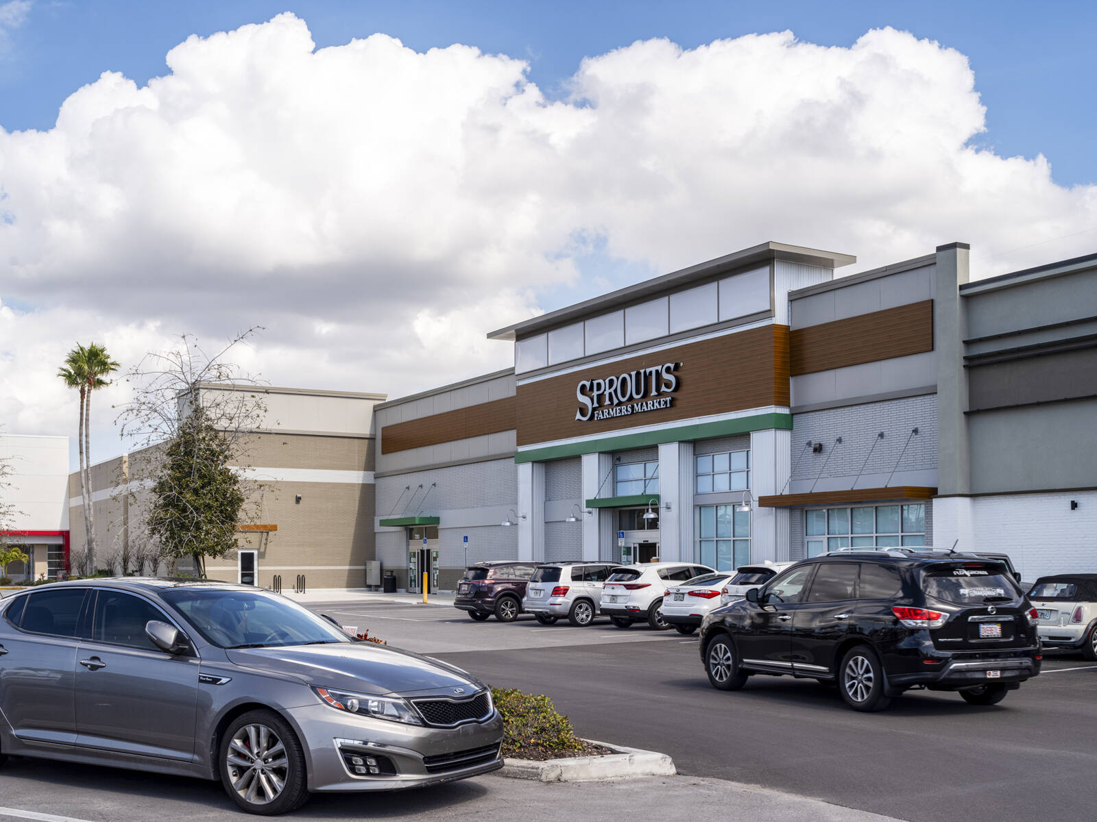 Silver car parked across from Sprouts Farmers Market entrance and black SUV driving by