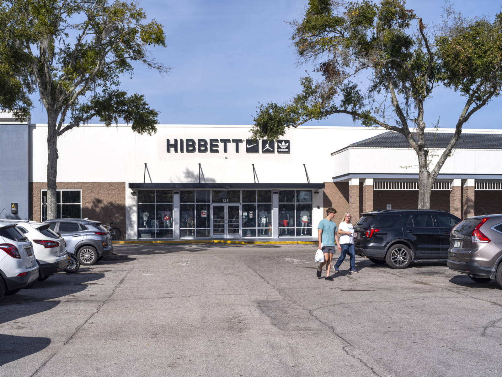 Hibbett sporting good store with trees and customers in parking aisle.