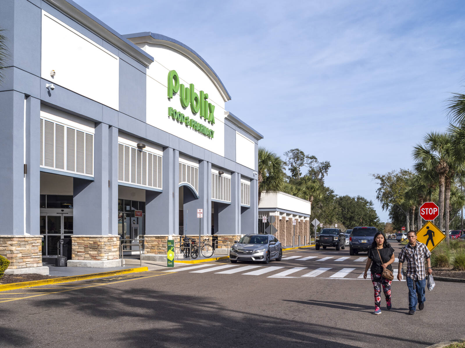 Two people at crosswalk of Publix with cars and palm trees behind them.