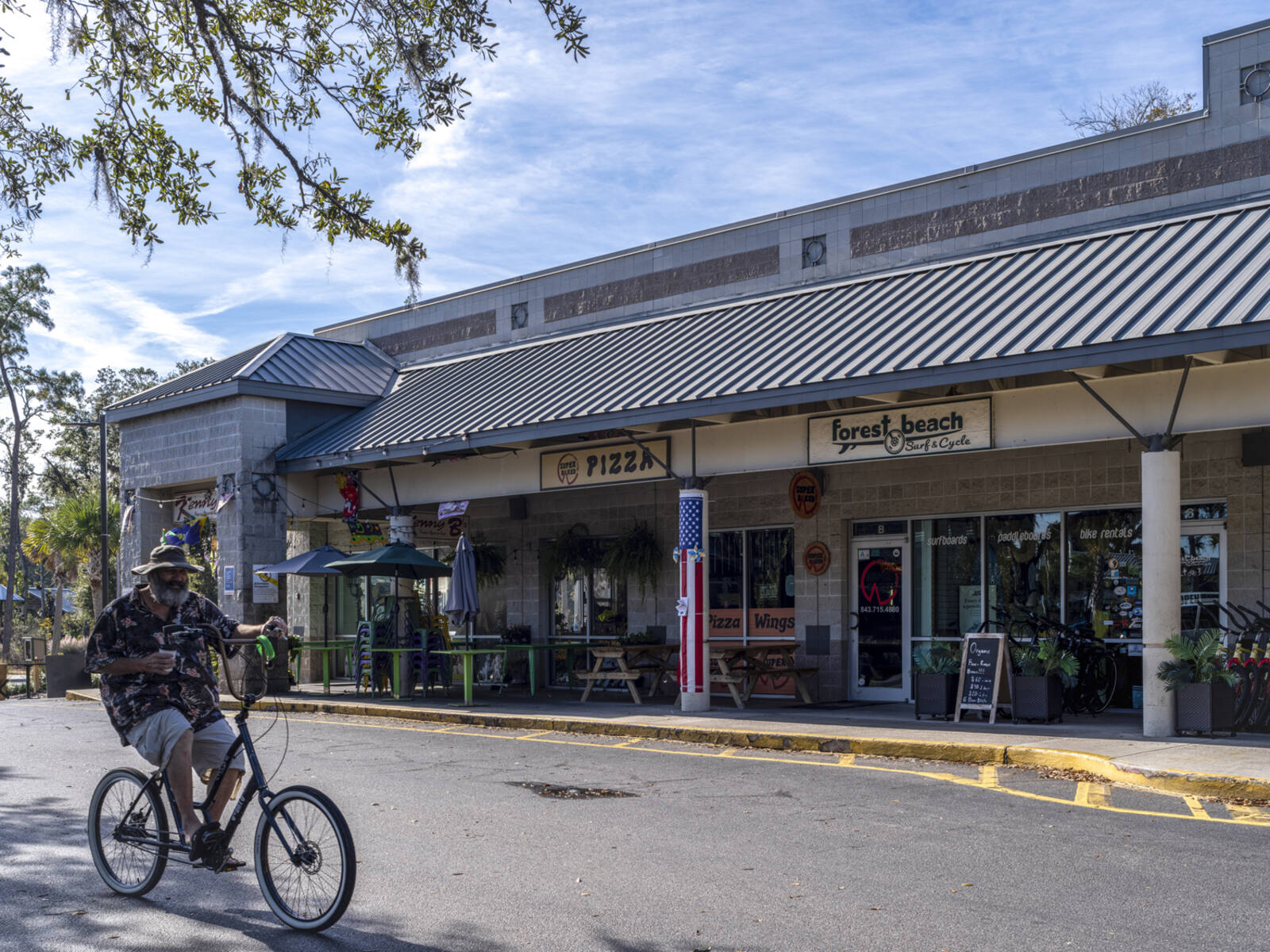 Man on bike riding past Pizza and Surf & Cycle shop at shopping center.