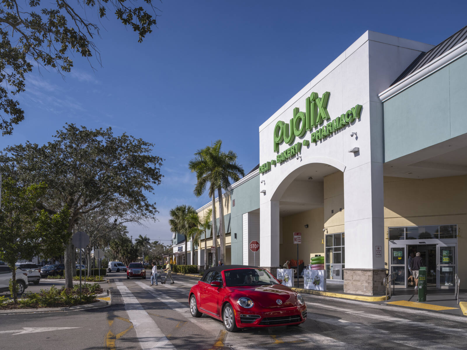 Red sports car passing Publix supermarket on tree-lined access road.