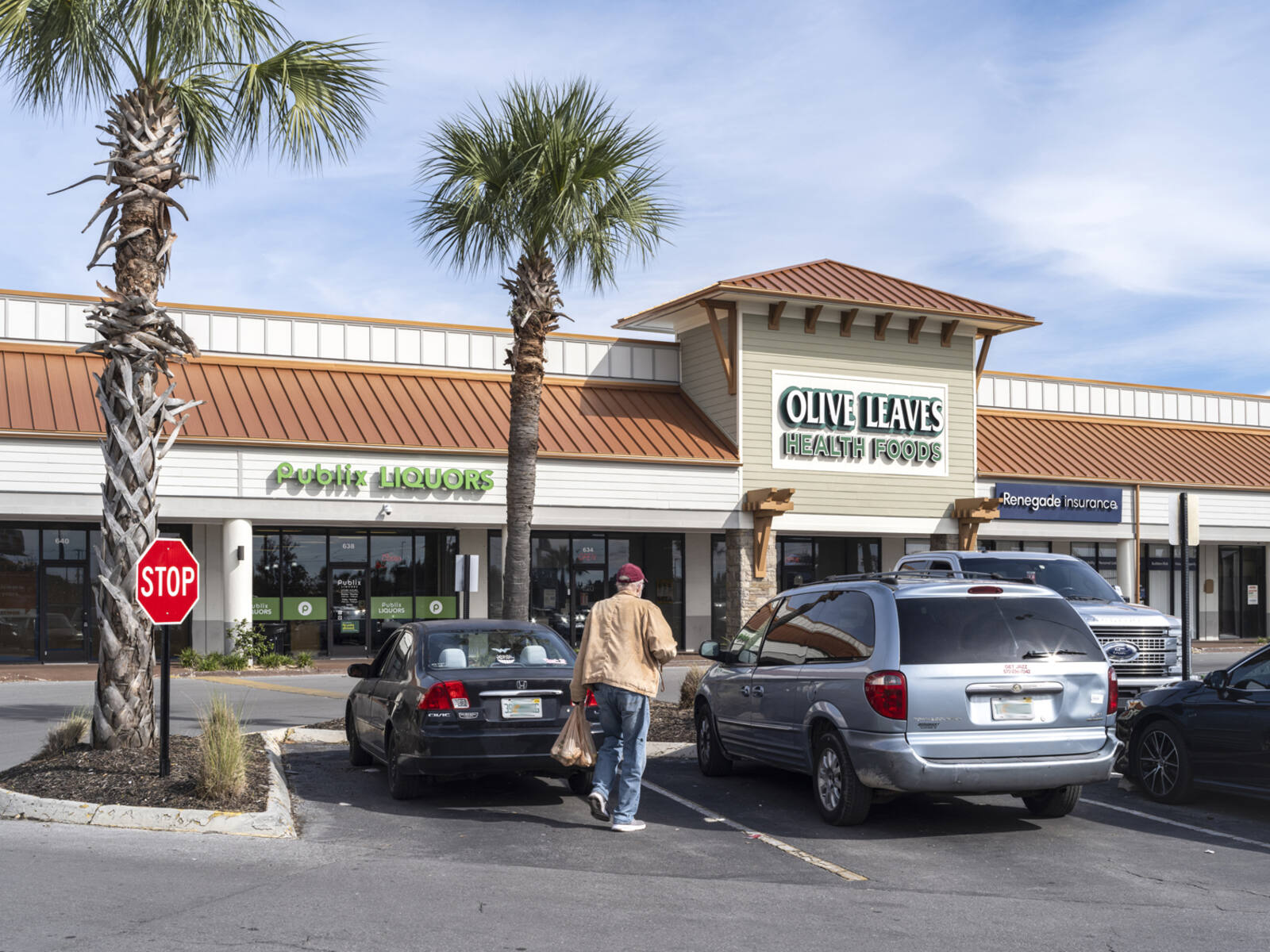 Man carrying bags in parking lot in front of Publix Liquors and Olive Leaves Health Foods.