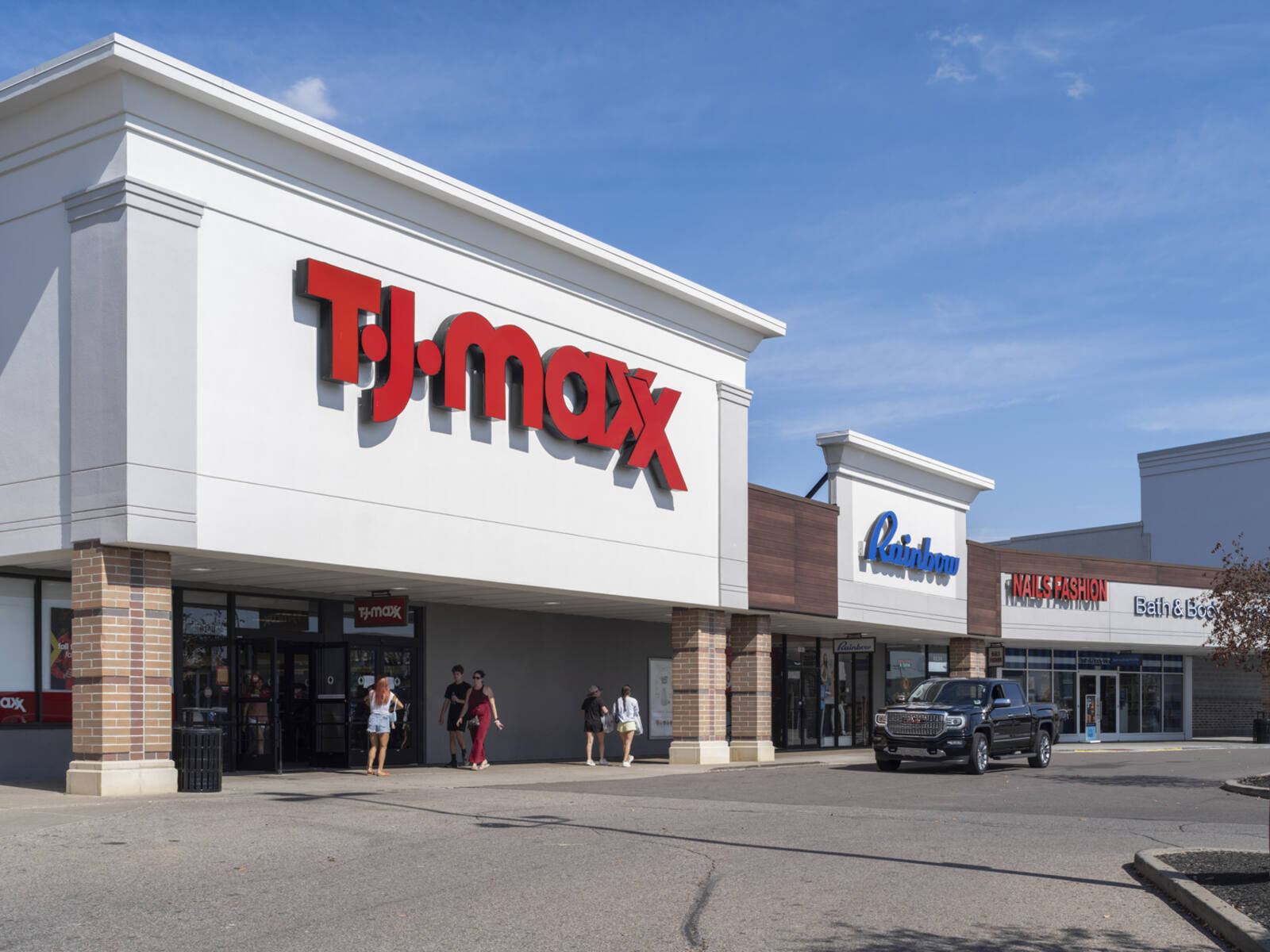 Black car approaches TJ Maxx on access road in shopping center.