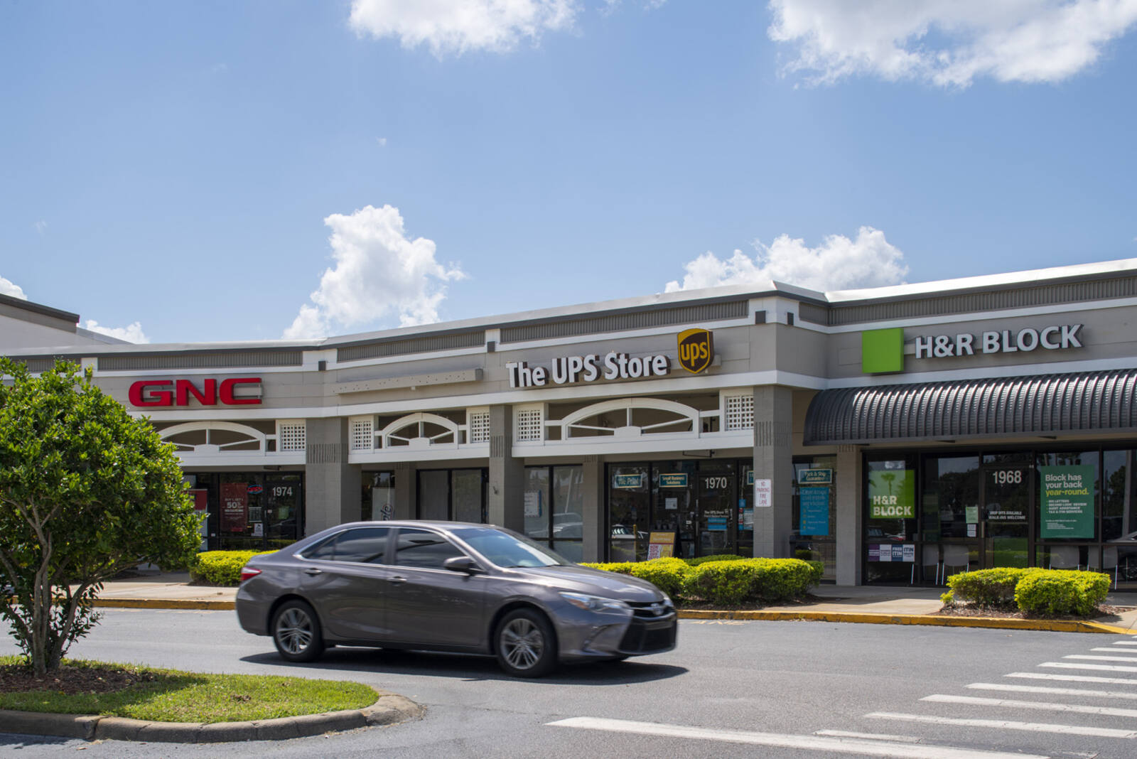 Grey car passing by front facades of GNC, UPS Store, H&R Block