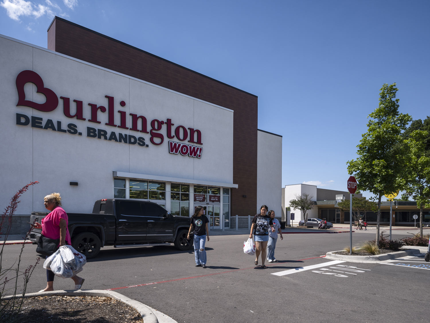 Shoppers with bags exiting Burlington store into crosswalk with truck and tree on right.