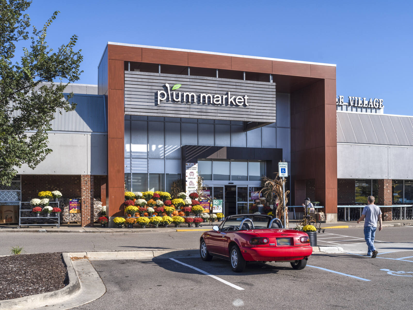 Red convertible parked in front of Plum Market with man walking towards entrance.