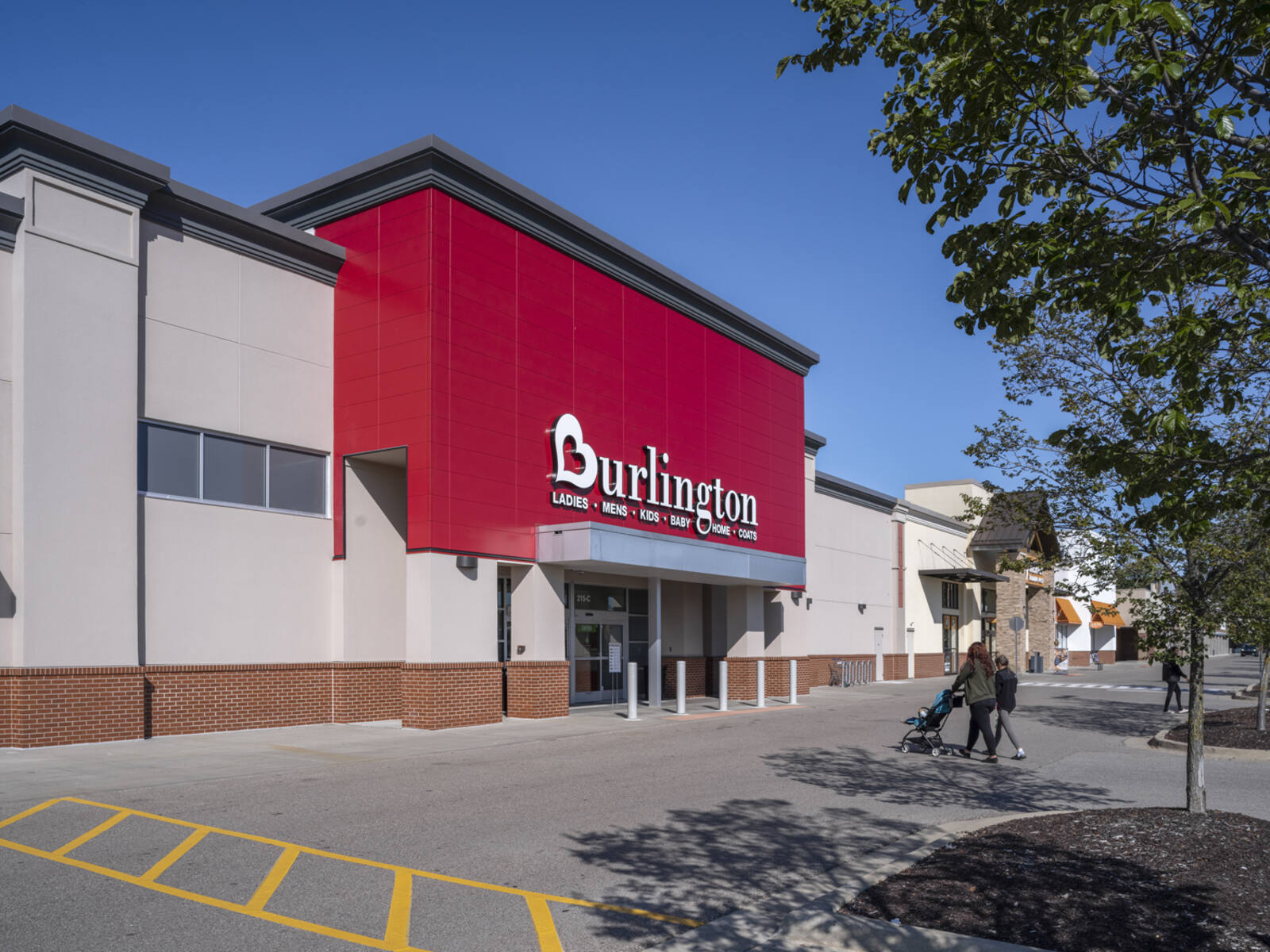 Woman with stroller and daughter approaching Burlington store with tree and planter in foreground.