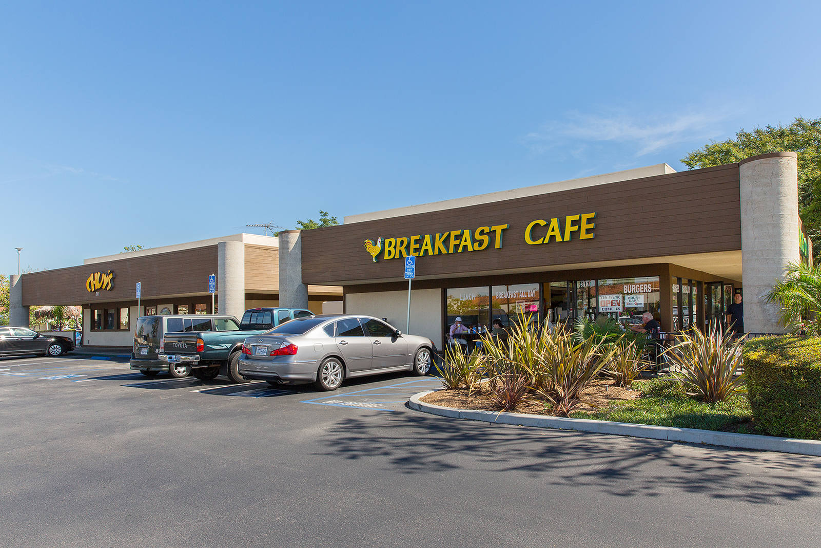 Cars lined in front of Breakfast Café with planters and bushes.