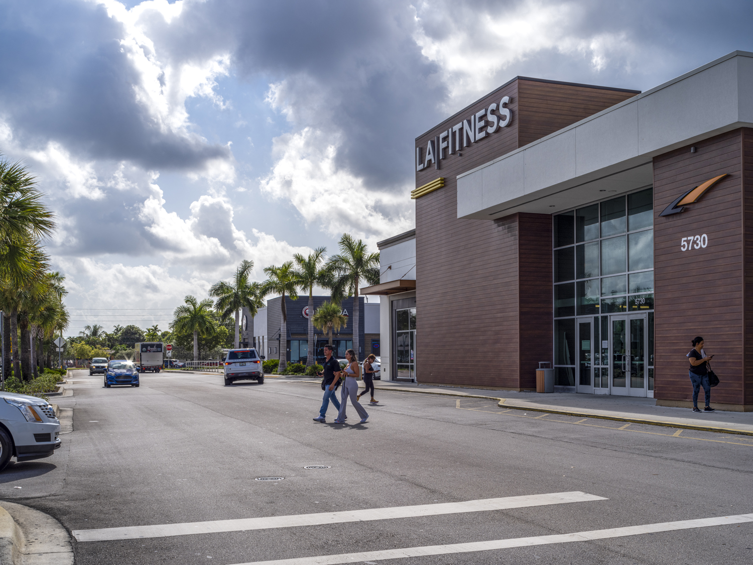 People at entrance of LA Fitness with row of palm trees behind.