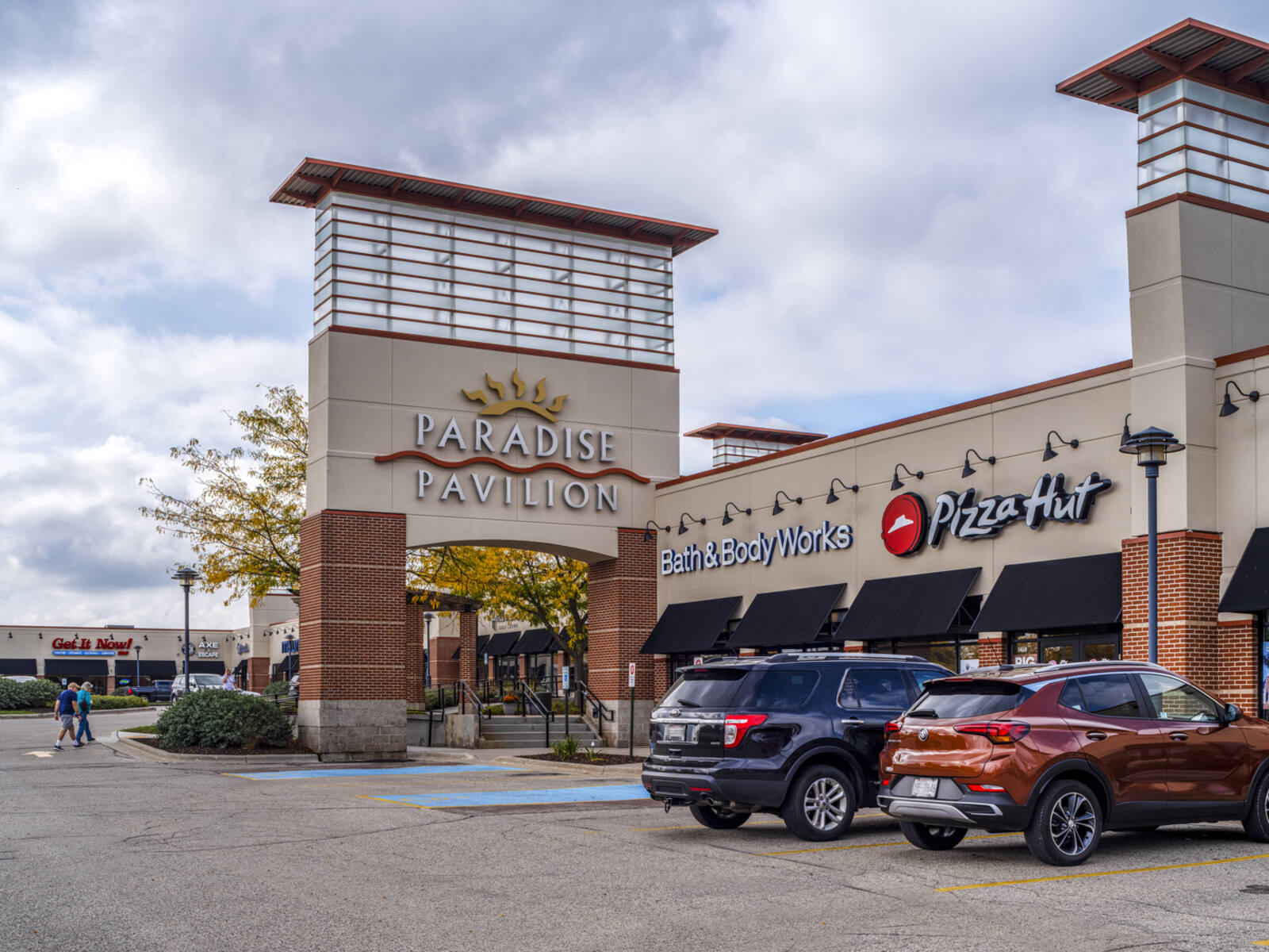 Paradise Pavilion sign on large pillar facing Pizza Hut and parked cars.