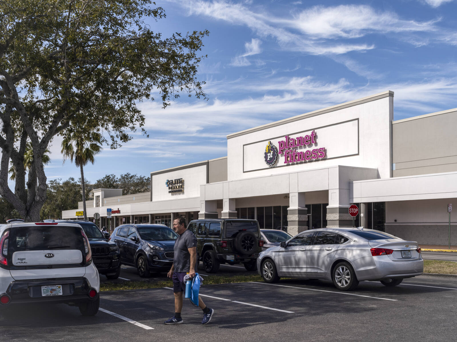Busy parking lot in front of Planet Fitness with man holding bag walking in foreground.