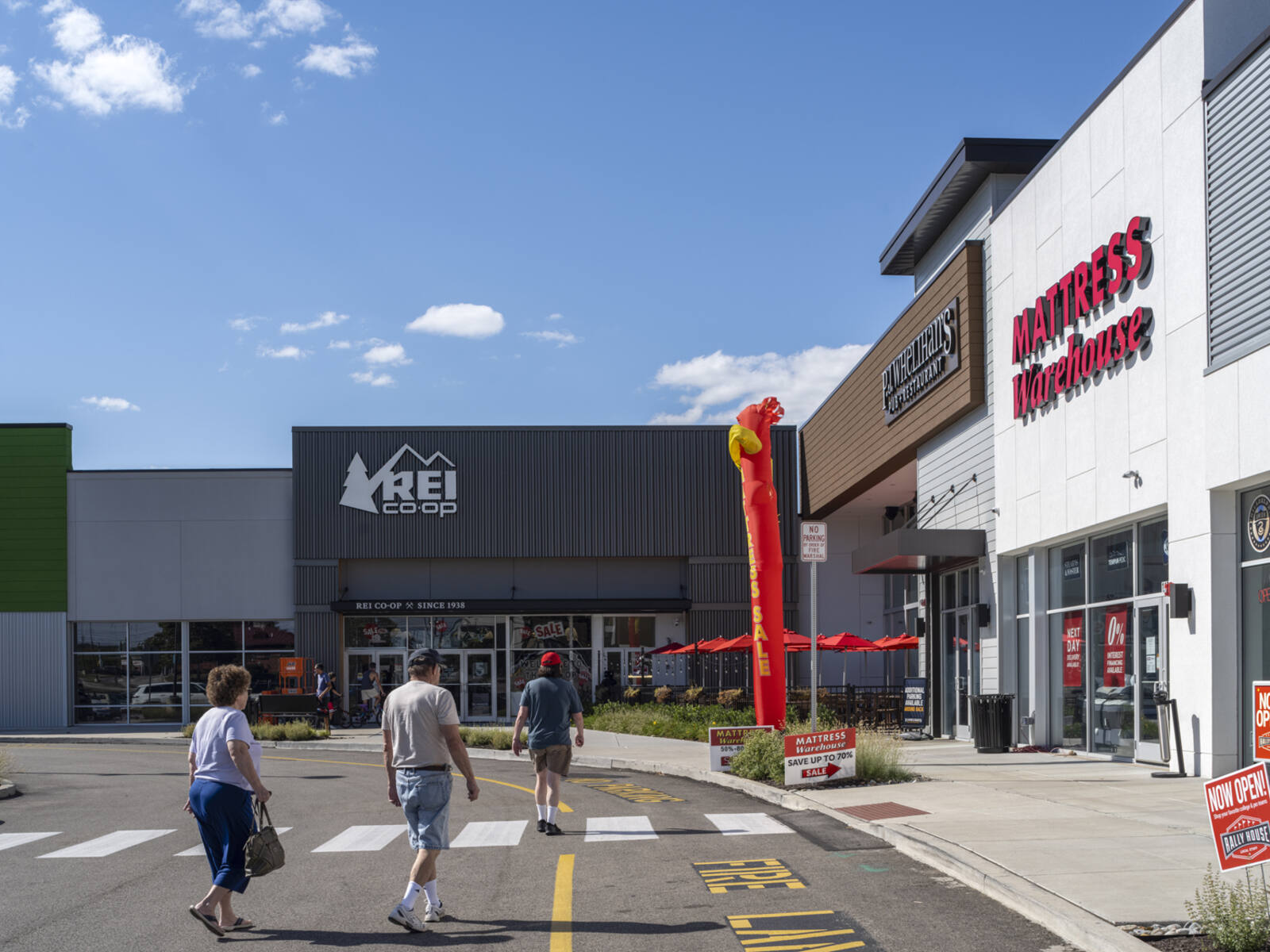 3 people approach REI store with Mattress Warehouse on the right.