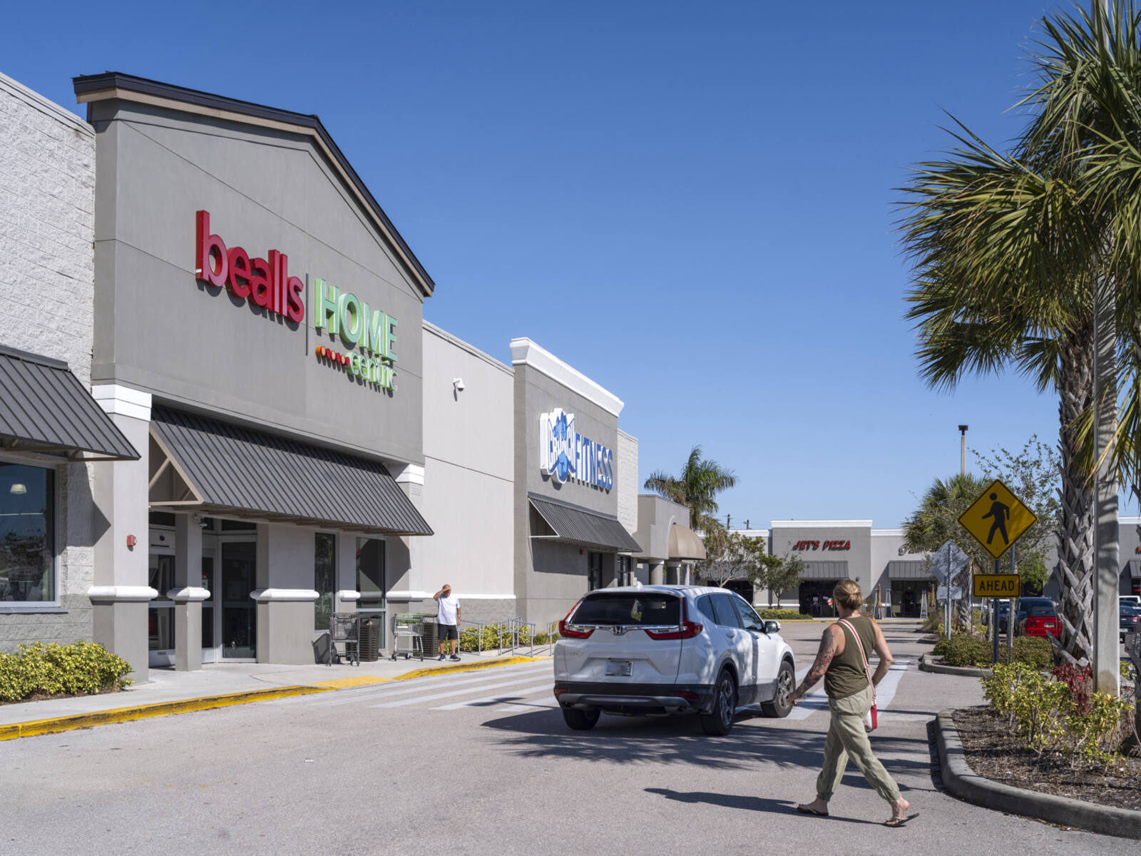 Woman crossing street with white SUV and walking towards bealls Home Centric store.