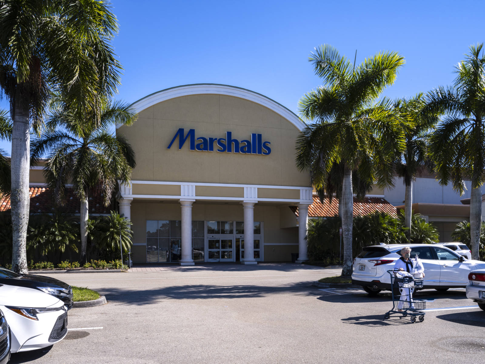 Shopper with cart leaving Marshalls through palm tree covered parking lot in Naples, FL.