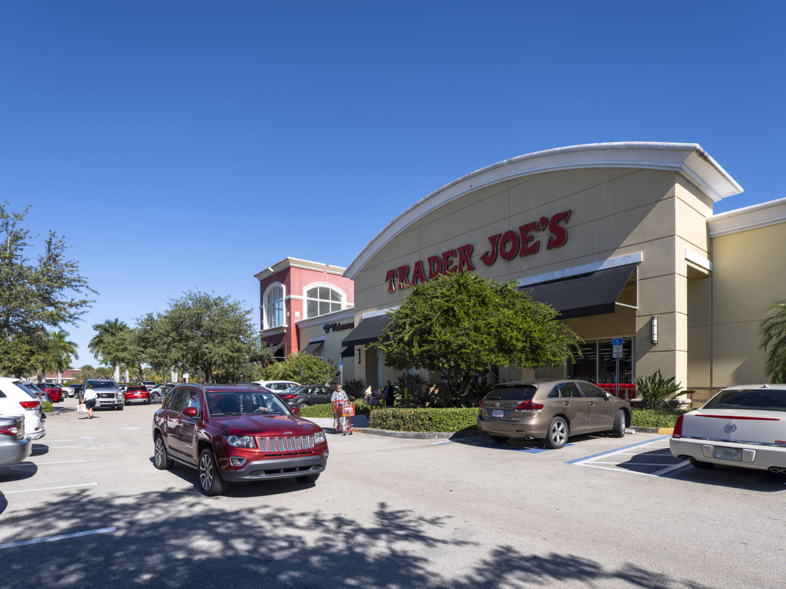 Trader Joe's with many trees in front and a red passing SUV at Granada Shoppes.