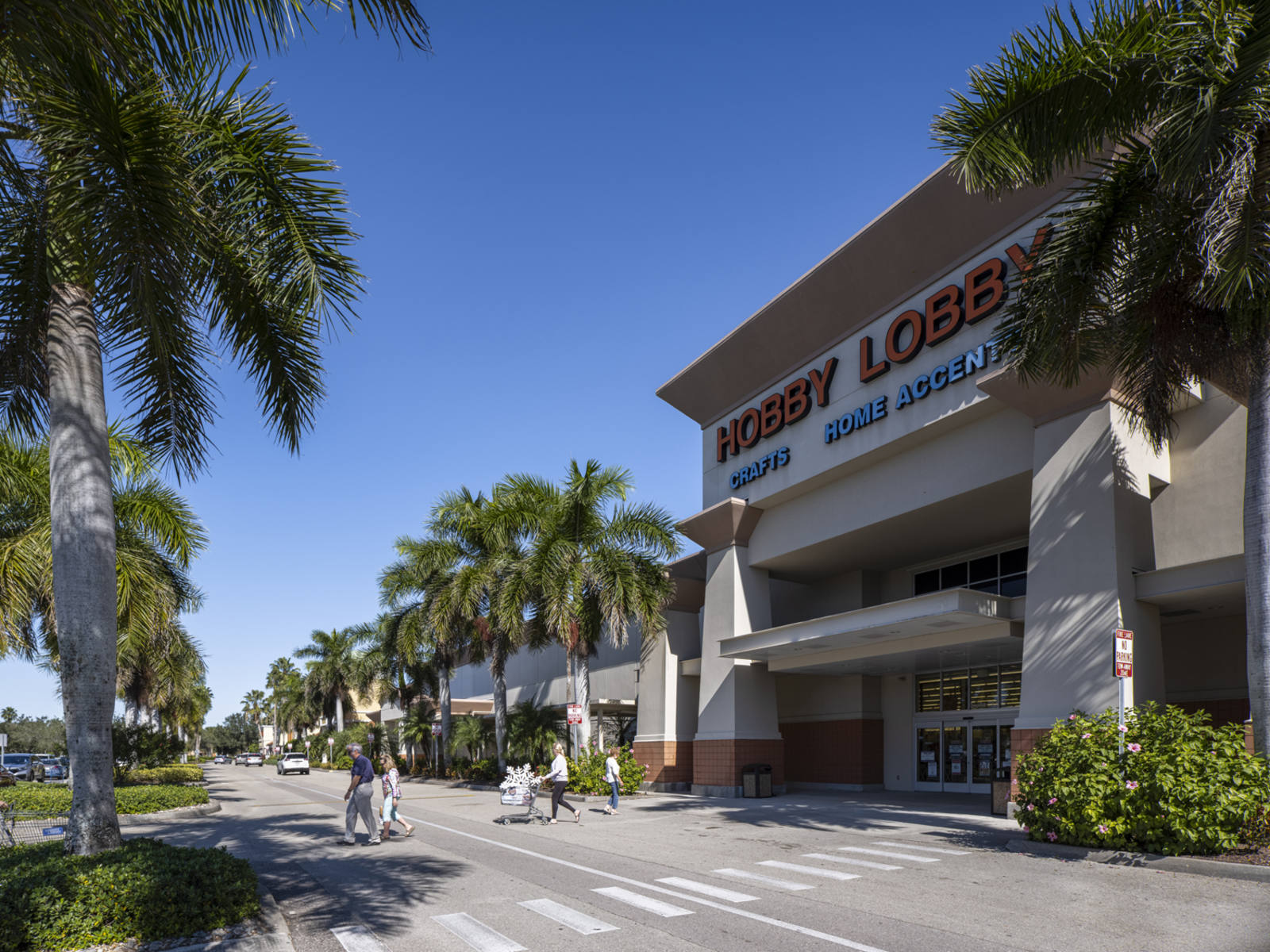 Customers with carts in Hobby Lobby craft store crosswalk with rows of palm trees.