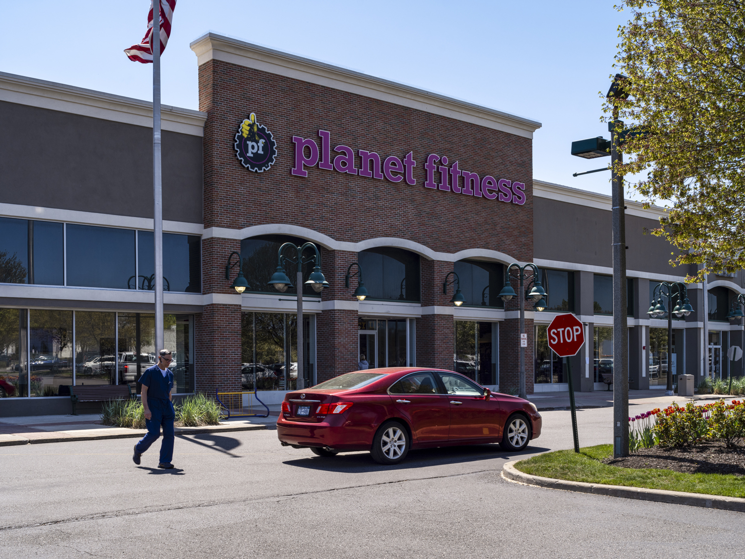 Man and red car in front of Planet Fitness gym with tree on the right.