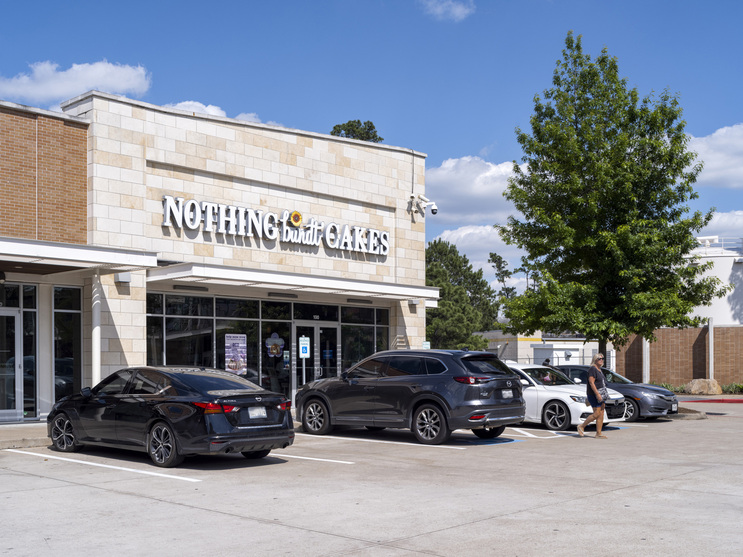 Nothing Bundt Cakes with large tree on right and customers and cars in parking lot.