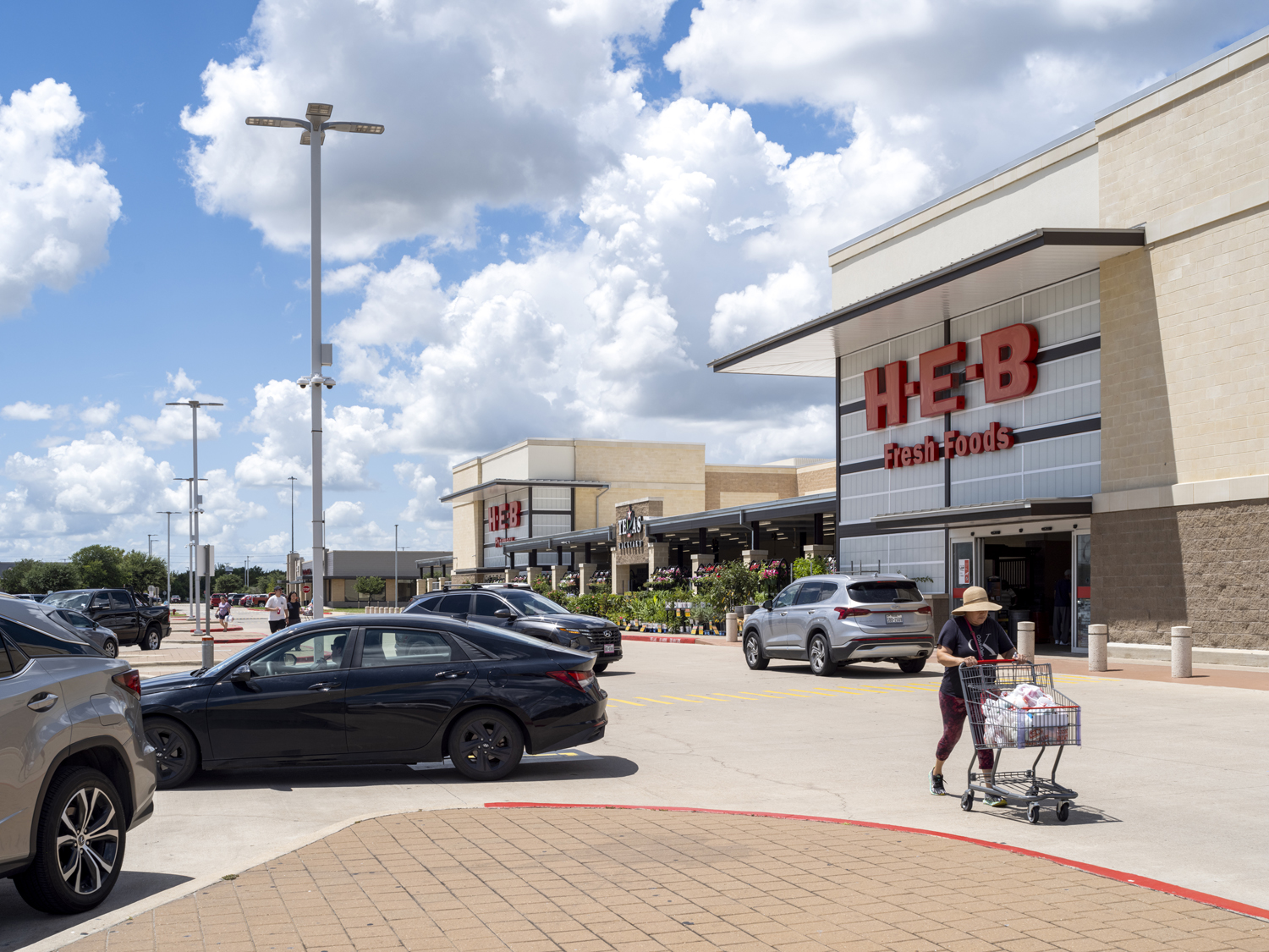 Woman with cart on road in front of busy HEB grocery store.