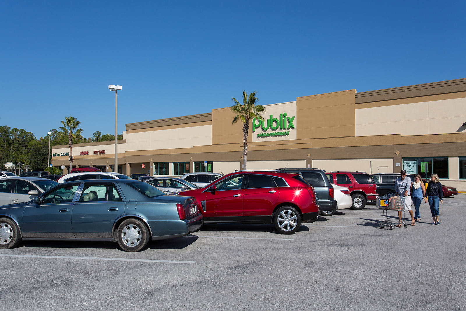 Shoppers with cart walking along parked cars at Publix supermarket lot.