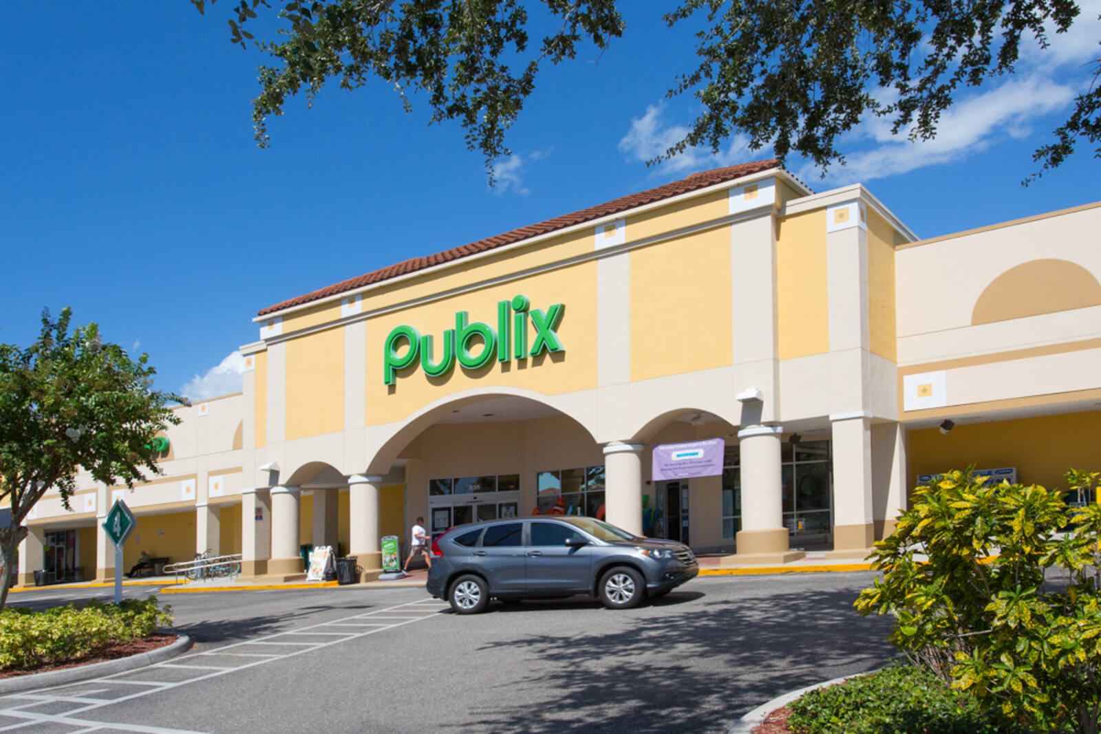 Grey SUV passing by entrance of Publix with trees on either side