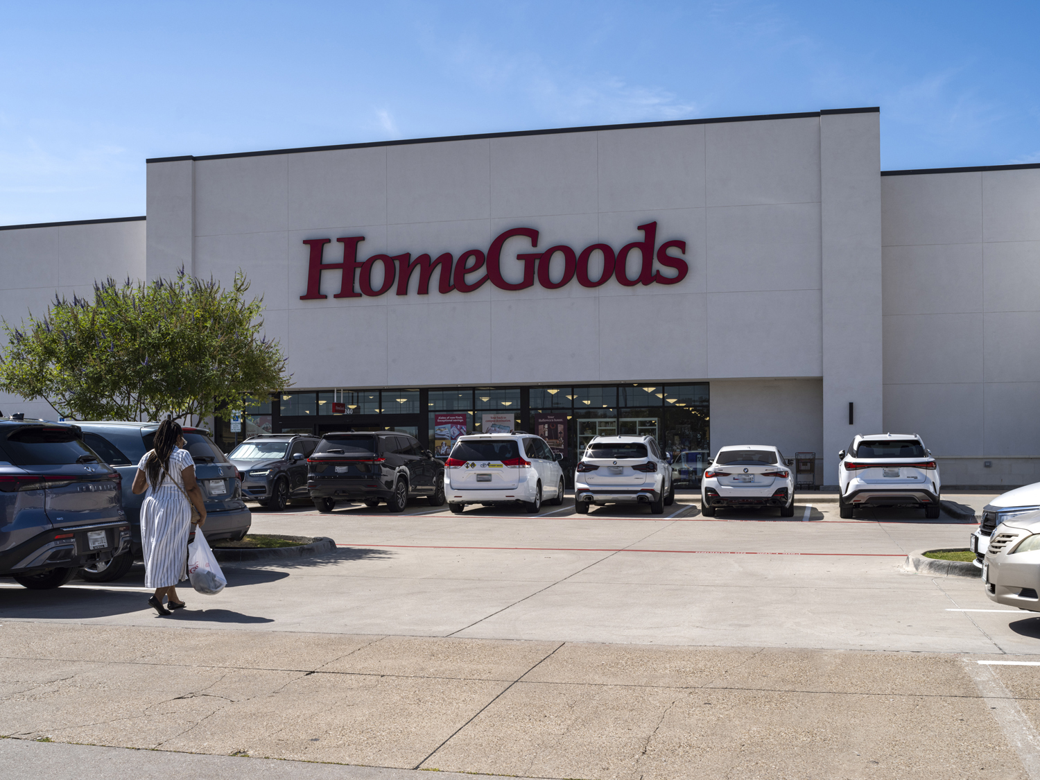 Woman approaching HomeGoods through parking lot with tree on the left.
