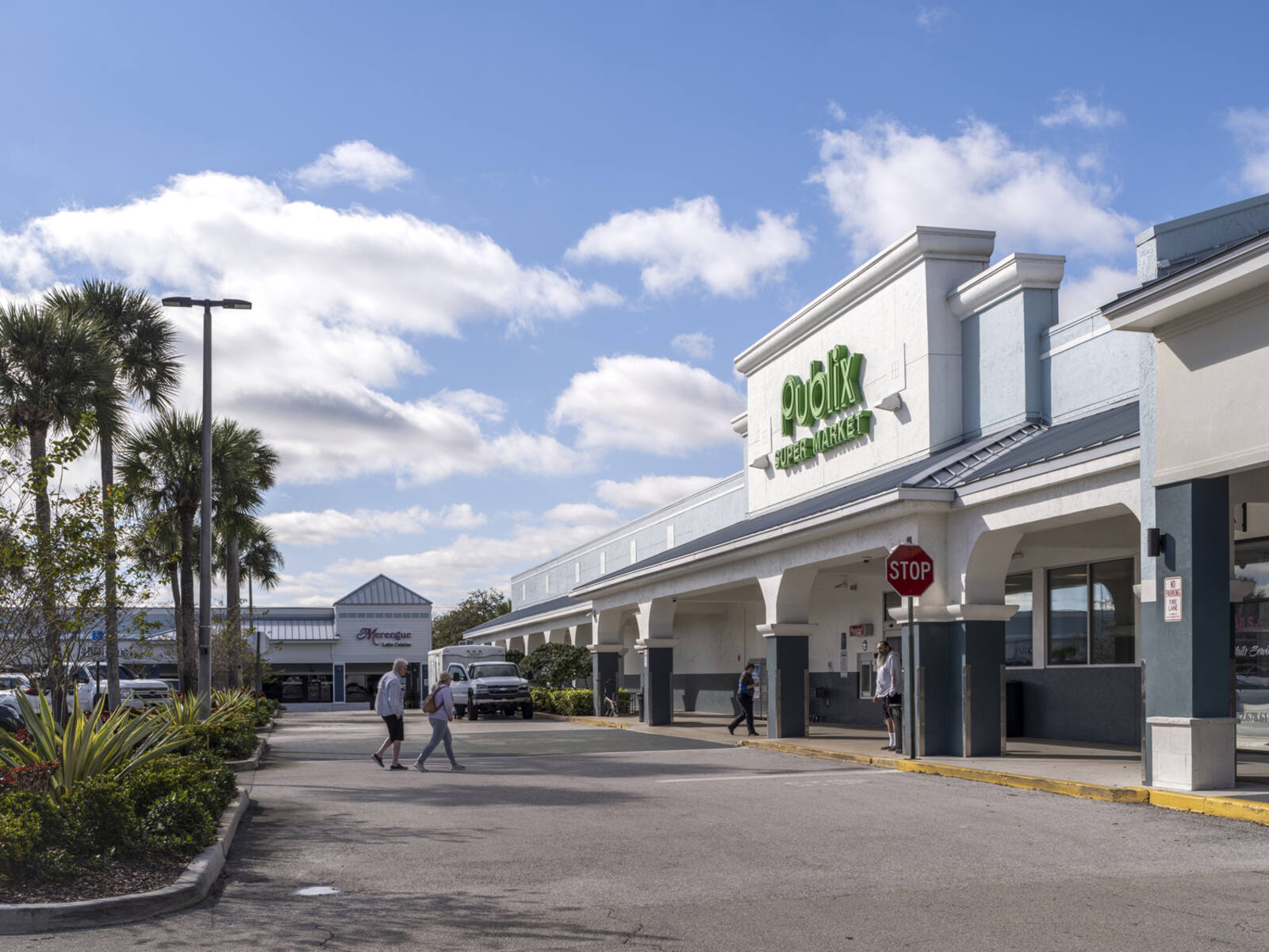 2 pedestrians walking towards Publix grocer with palm trees on left.