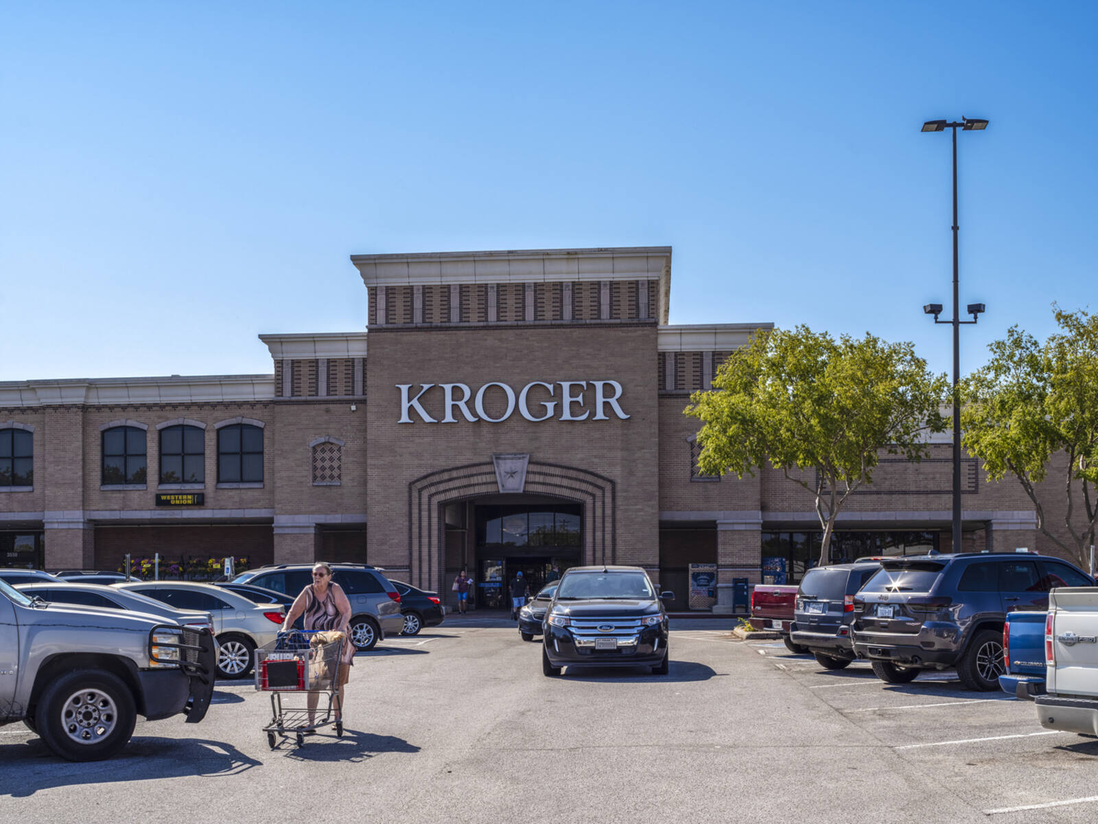 Shopper with cart walking up aisle in busy parking lot of Kroger supermarket.