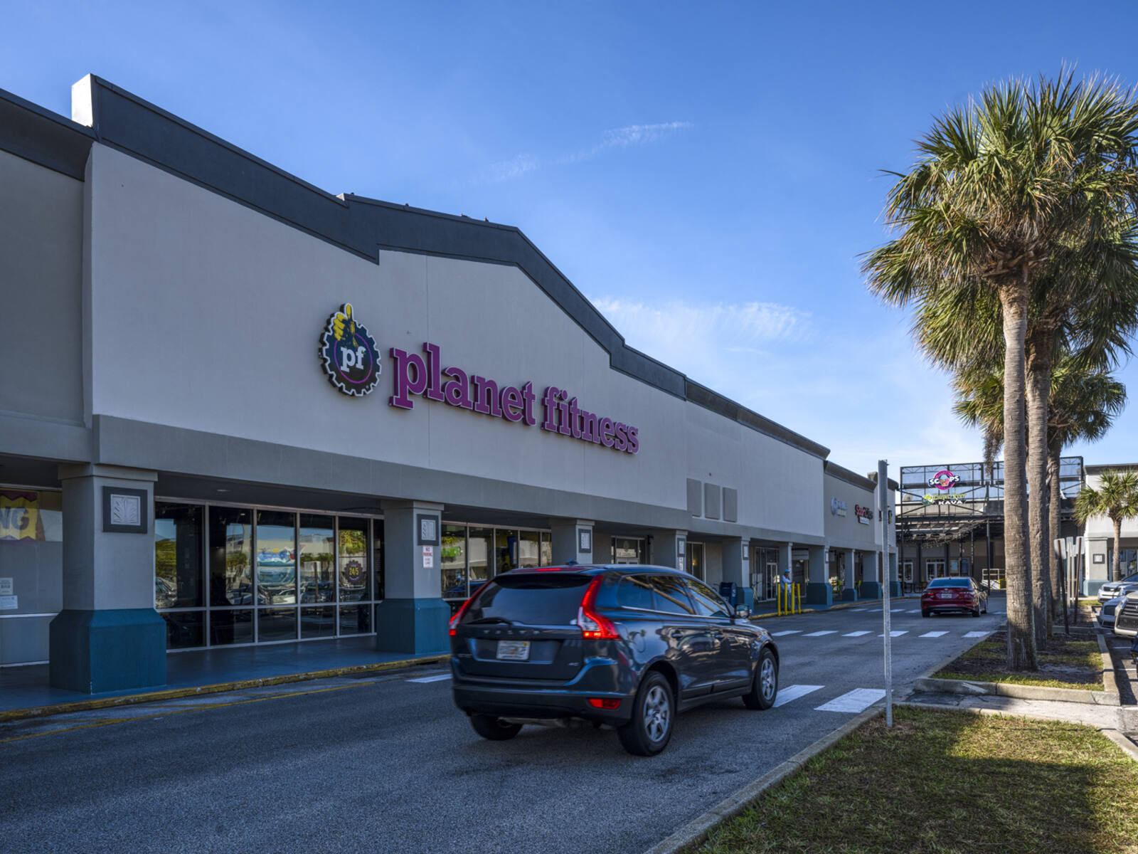 Dark SUV passes Planet Fitness on palm tree-lined access road.