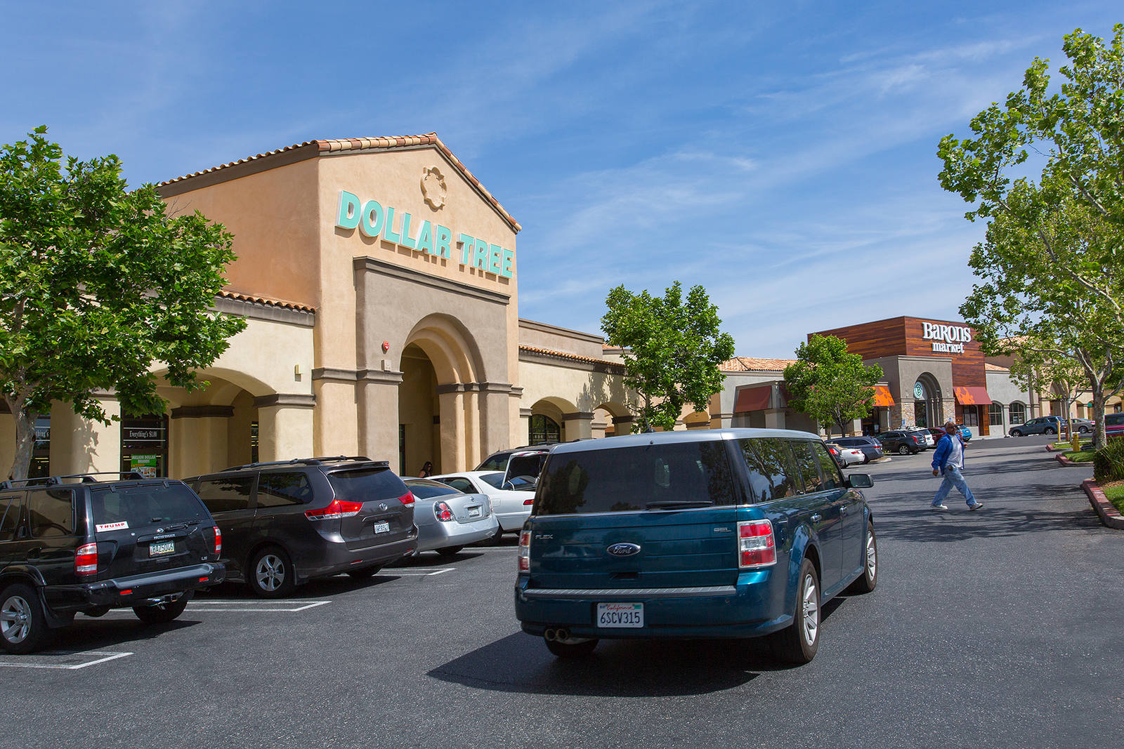 Green minivan passes rows of trees and cars in Dollar Tree parking lot.