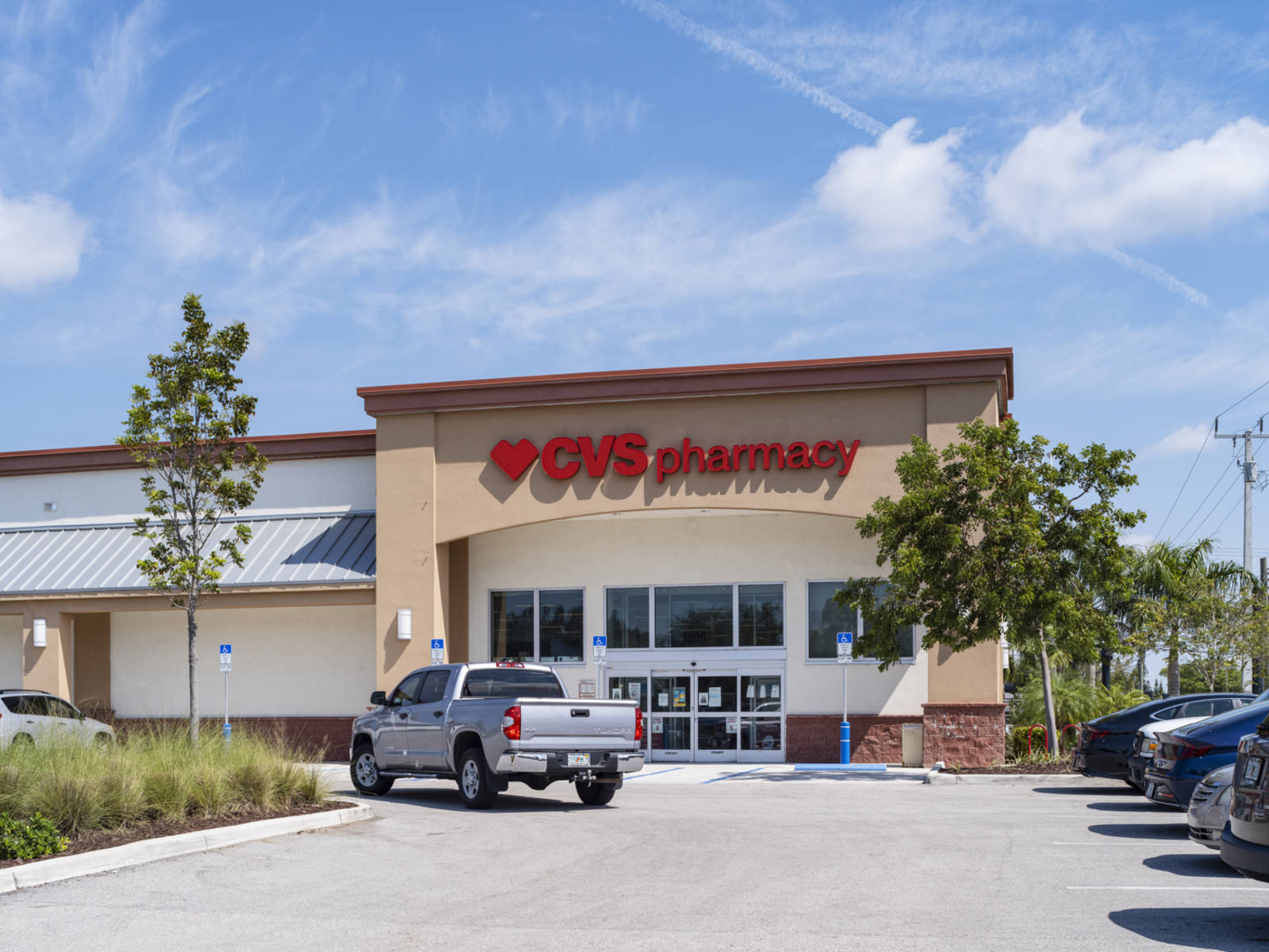 Grey truck driving past CVS Pharmacy entrance with trees.