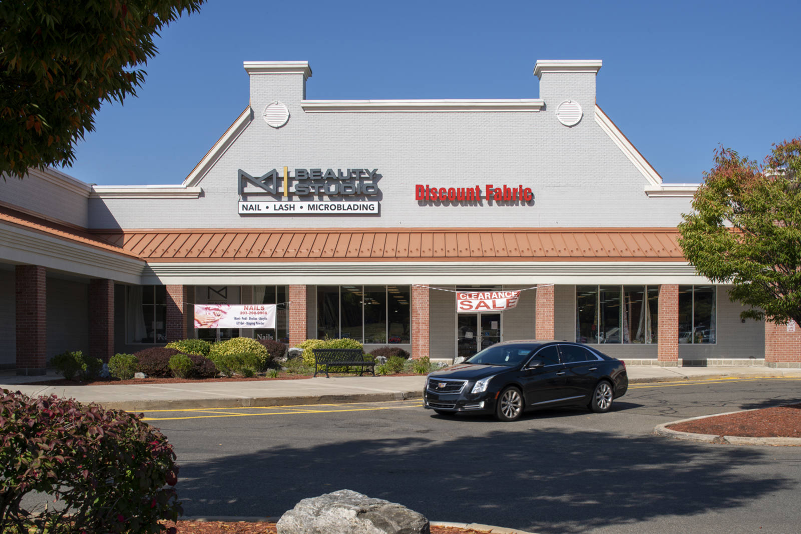 Black sedan driving by Beauty Studio and Discount Fabrics in tree-lined shopping center.