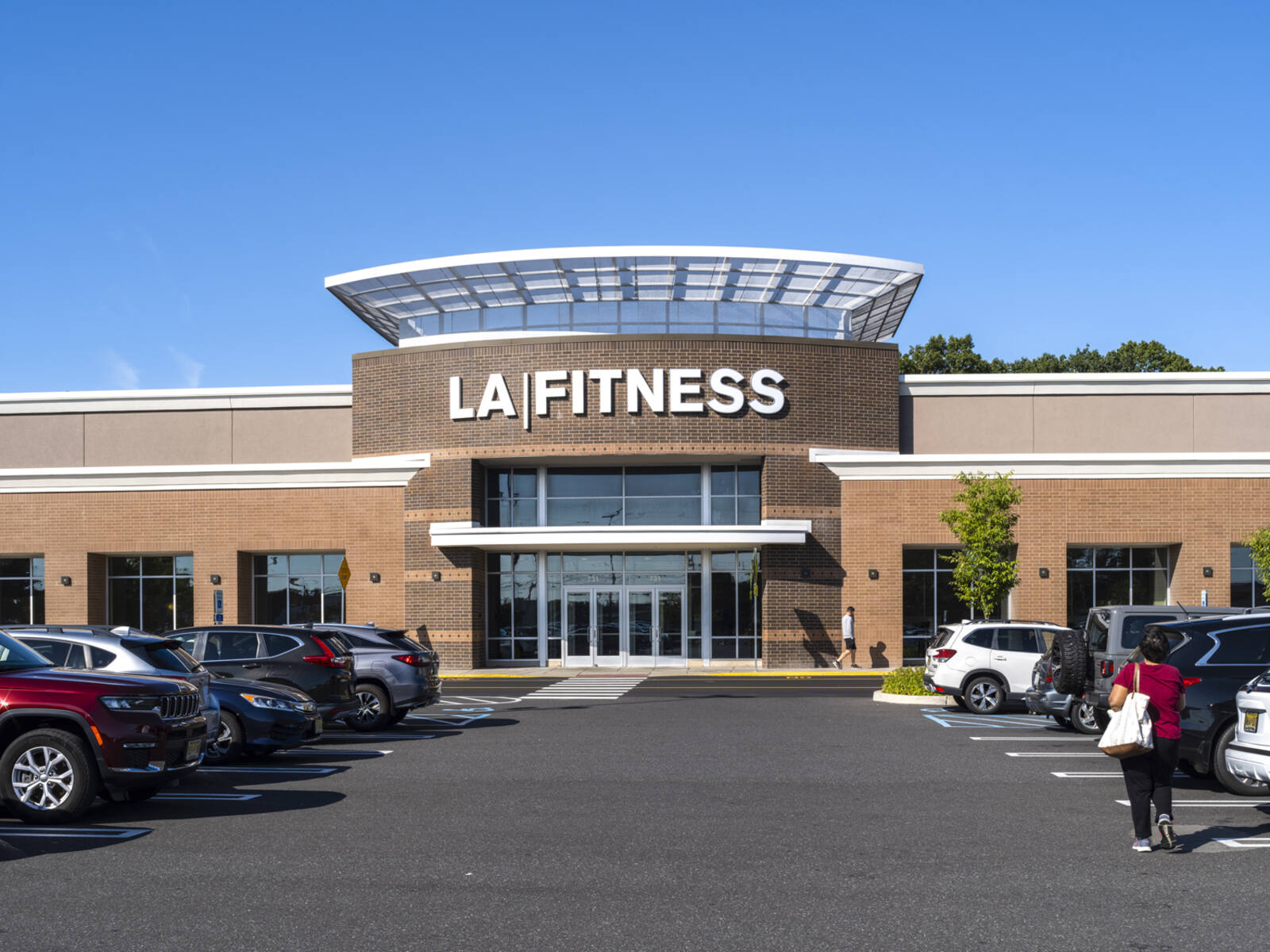Woman approaches LA Fitness via an aisle in busy parking lot.
