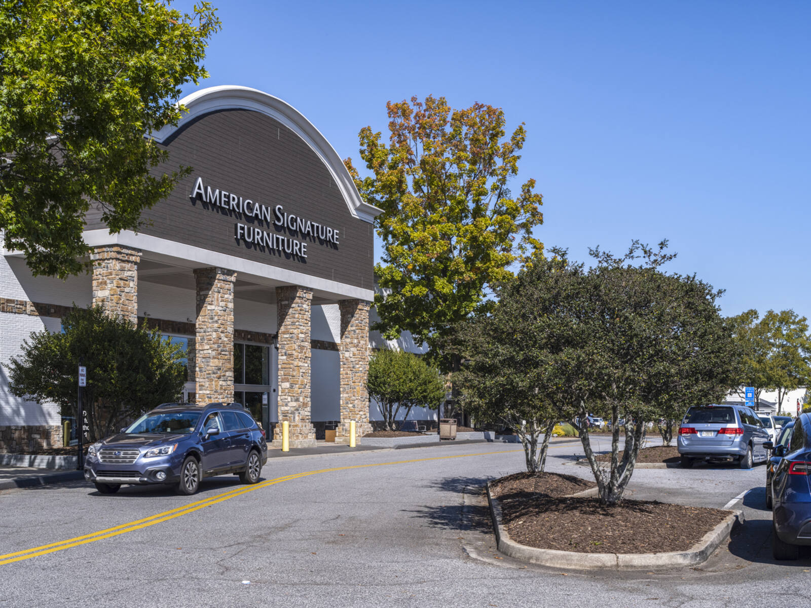 SUV passes American Signature Furniture with trees on both sides of road.