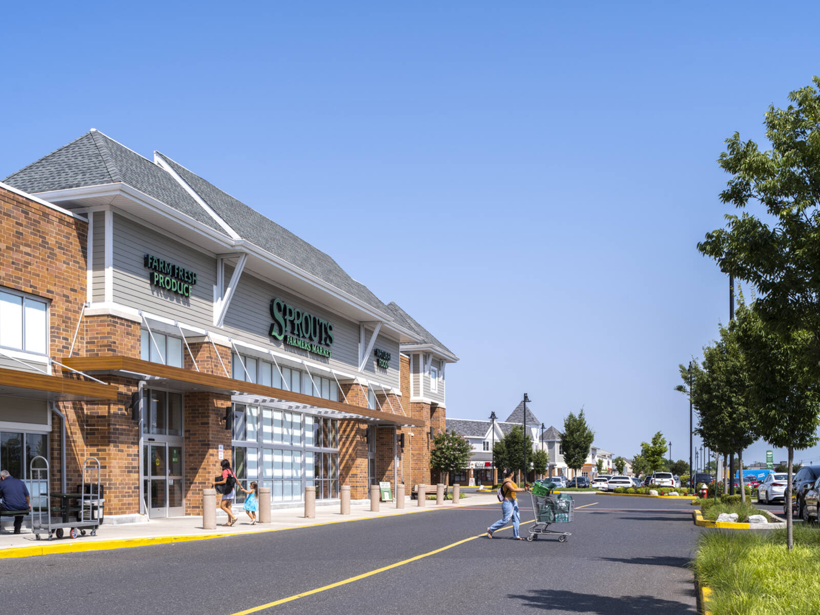 Patrons enter and exit Sprouts Farmers Market in crosswalk with trees and grass.
